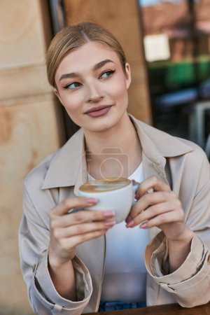 pretty blonde woman in trench coat enjoying her cup of cappuccino in cafe, relaxed atmosphere
