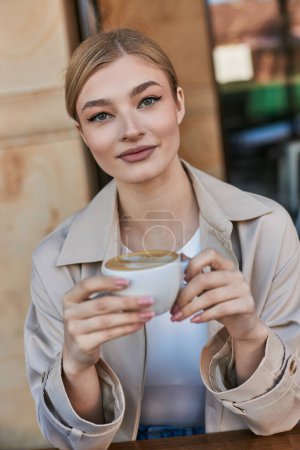 joven mujer rubia en gabardina disfrutando de su taza de capuchino en la cafetería, ambiente relajado
