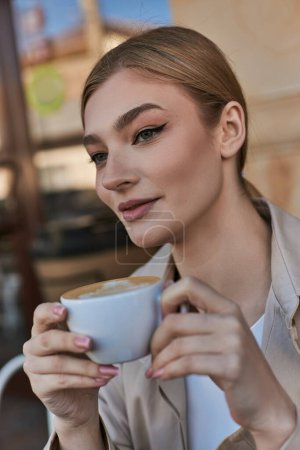 elegant blonde woman in trench coat enjoying her cup of cappuccino in cafe, relaxed atmosphere