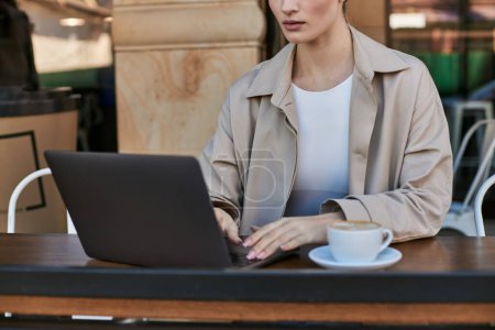 Photo for Cropped view of young woman in trench coat using laptop near cup of cappuccino in cafe, remote work - Royalty Free Image