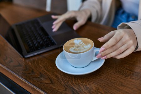 Photo for Cropped shot of young woman in trench coat using laptop near cup of cappuccino in cafe, remote work - Royalty Free Image