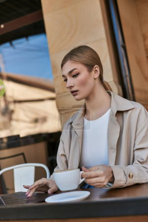 Photo for Blonde young woman in trench coat using laptop near cup of cappuccino in cafe, remote work - Royalty Free Image