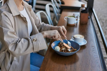 abgeschnittene Ansicht einer Frau im Trenchcoat, die belgische Waffeln mit Eis neben einer Tasse Kaffee isst