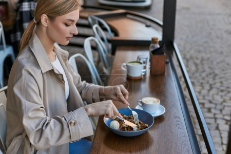 blonde young woman in trench coat eating her belgian waffles with ice cream next to cup of coffee