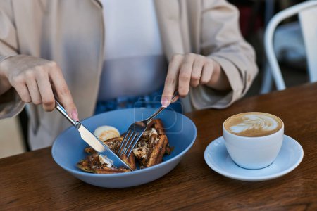 cropped view of woman in trench coat eating belgian waffles with ice cream next to cup of coffee