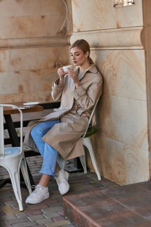 atractiva mujer joven con elegante gabardina sentado y sosteniendo la taza de café dentro de la cafetería