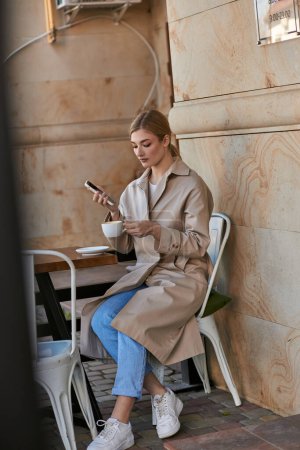 atractiva mujer joven con elegante gabardina usando teléfono inteligente y sosteniendo la taza de café en la cafetería