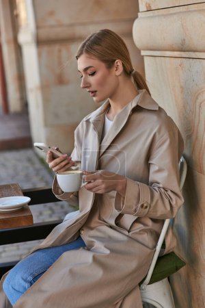 atractiva mujer joven con elegante gabardina usando teléfono inteligente cerca de la taza de café en la mesa en la cafetería