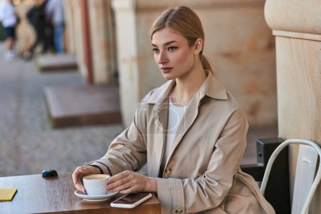 mujer joven rubia en gabardina sentada cerca de la taza de café y teléfono inteligente en la mesa en la cafetería