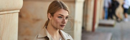 young woman with blonde hair in stylish trench coat looking away outdoors in cafe, banner
