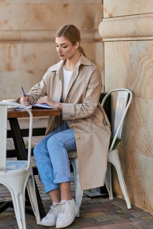 Blonde junge Frau im Trenchcoat macht sich Notizen in Notizbuch neben Kaffeetasse im Café, Planungstag