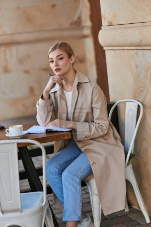 Nachdenkliche blonde Frau im Trenchcoat sitzt am Tisch und hält Stift am Kinn, schreibt Tagebuch