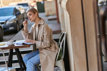 nachdenkliche Frau im Trenchcoat, die mit einer Tasse Kaffee am Tisch sitzt und Stift in der Hand hält, Tagebuch schreibt
