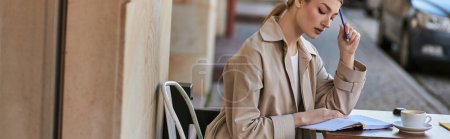 woman in trench coat sitting near cup of coffee and holding pen near notebook, writing diary banner