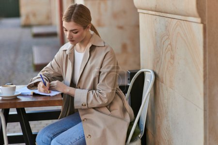 pensive young woman in stylish trench coat writing in notebook near cup of coffee, diary