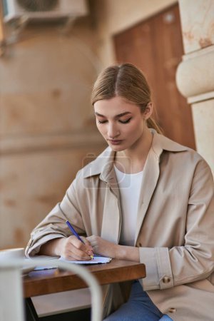 pretty young woman in stylish trench coat writing in notebook near cup of coffee, taking notes