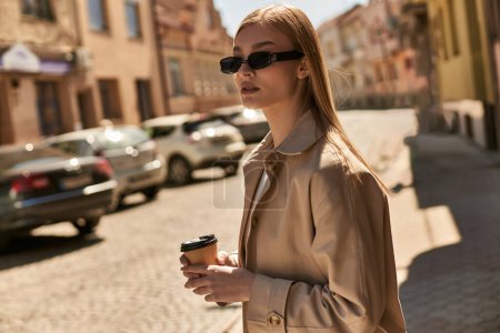 blonde woman in stylish trench coat holding coffee to go while walking on street in sunny day