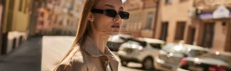 young blonde woman in stylish trench coat and sunglasses walking on street in sunny day, banner