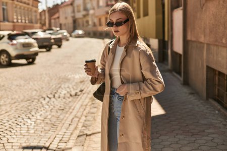 blonde woman in trendy trench coat holding coffee to go while walking on street in sunny day
