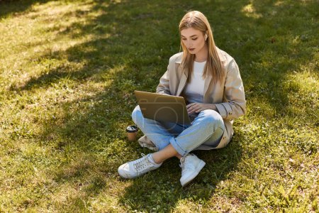 femme blonde dans les écouteurs et trench coat assis sur l'herbe près de tasse en papier et en utilisant un ordinateur portable