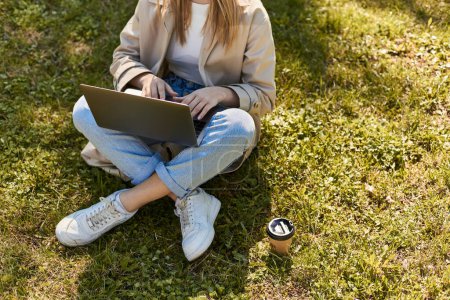 gestutzte Frau in Jeans und Trenchcoat sitzt auf grünem Rasen neben Pappbecher und benutzt Laptop