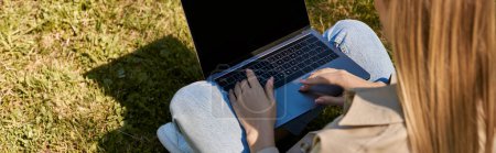 vue aérienne, femme en jeans et trench coat assis sur la pelouse verte et à l'aide d'un ordinateur portable, bannière