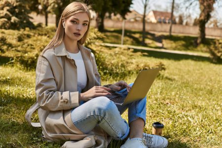 femme blonde dans les écouteurs et trench manteau à l'aide d'un ordinateur portable tout en étant assis sur l'herbe près de tasse en papier