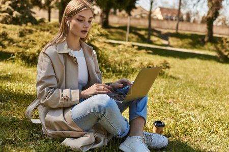 blonde jeune femme dans les écouteurs et trench coat en utilisant un ordinateur portable tout en étant assis sur l'herbe près de tasse de papier