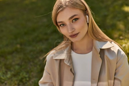 portrait of pretty young woman in wireless earphones and beige trench coat looking at camera in park