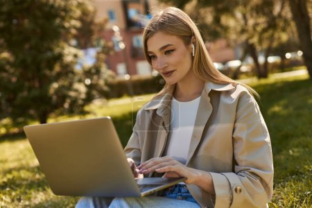 attractive young woman in wireless earphones and trench coat using laptop while sitting on grass