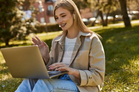 happy young woman in wireless earphones and trench coat using laptop while having video call