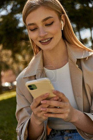 glückliche blonde Frau in kabellosen Kopfhörern und beigem Trenchcoat mit ihrem Smartphone im Park