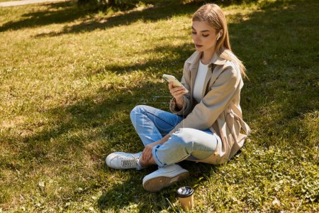 junge blonde Frau in drahtlosen Kopfhörern und Trenchcoat, die ihr Smartphone benutzt und auf dem Rasen sitzt