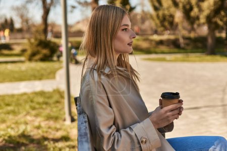 Photo for Young blonde woman in trench coat holding paper cup with takeaway coffee, sitting on bench in park - Royalty Free Image