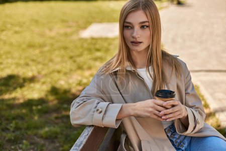 joven rubia en gabardina sosteniendo taza de papel con café y sentada en el banco en el parque