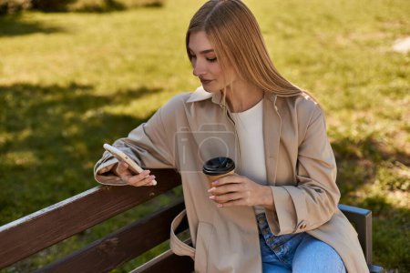 Blondine im Trenchcoat hält Pappbecher mit Kaffee und Smartphone in der Hand, sitzt auf Bank