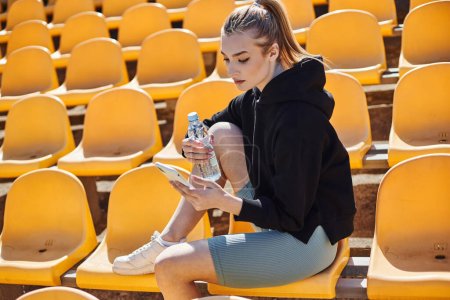 sporty woman with ponytail holding bottle of water and using smartphone after workout in stadium