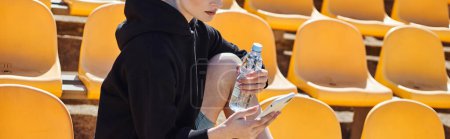 cropped sporty woman holding bottle of water and using smartphone after workout in stadium, banner