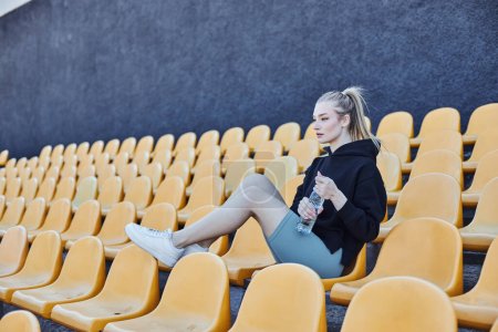 Sportlerin mit Pferdeschwanz hält Wasserflasche in der Hand und sitzt nach dem Training auf dem Stadionstuhl