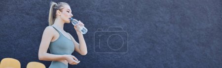Photo for Athletic blonde sportswoman in activewear drinking water and standing near stadium chairs, banner - Royalty Free Image