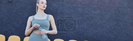 Photo for Athletic and blonde sportswoman in activewear holding bottle of water near stadium chairs, banner - Royalty Free Image