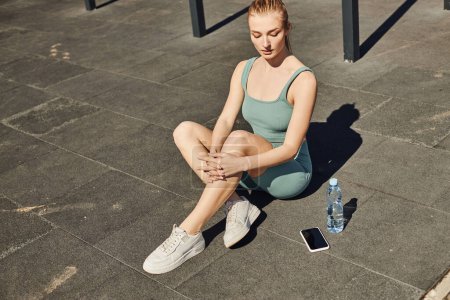 Blonde Sportlerin in enger Aktivkleidung sitzt neben Wasserflasche und Smartphone auf dem Boden