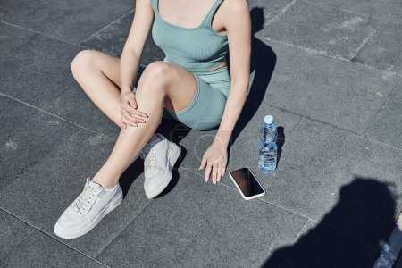 gestutzte Sportlerin in enger Aktivkleidung sitzt neben Wasserflasche und Smartphone auf dem Boden