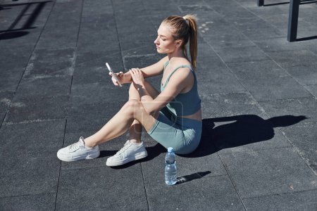 fitte Frau in enger Aktivkleidung, die neben einer Wasserflasche sitzt und nach dem Training das Smartphone benutzt