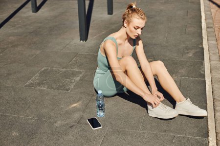 young fit woman in tight activewear sitting next to bottle of water and mobile phone after workout