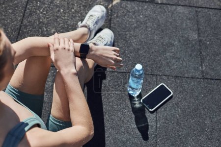 top view of fit woman in activewear checking fitness tracker next to smartphone and water bottle