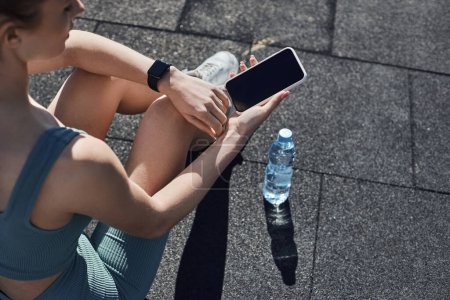 top view of fit woman in activewear with fitness tracker on wrist using smartphone near water bottle