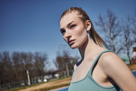 Porträt einer blonden und jungen Sportlerin in grauem Crop Top, die nach dem Training im Freien wegschaut