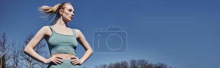 low angle view of sportswoman in activewear standing with hands on hips and looking away, banner