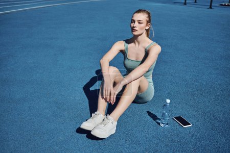 Foto de Mujer atlética joven en ropa deportiva sentada junto a la botella de agua y el teléfono móvil después del entrenamiento - Imagen libre de derechos